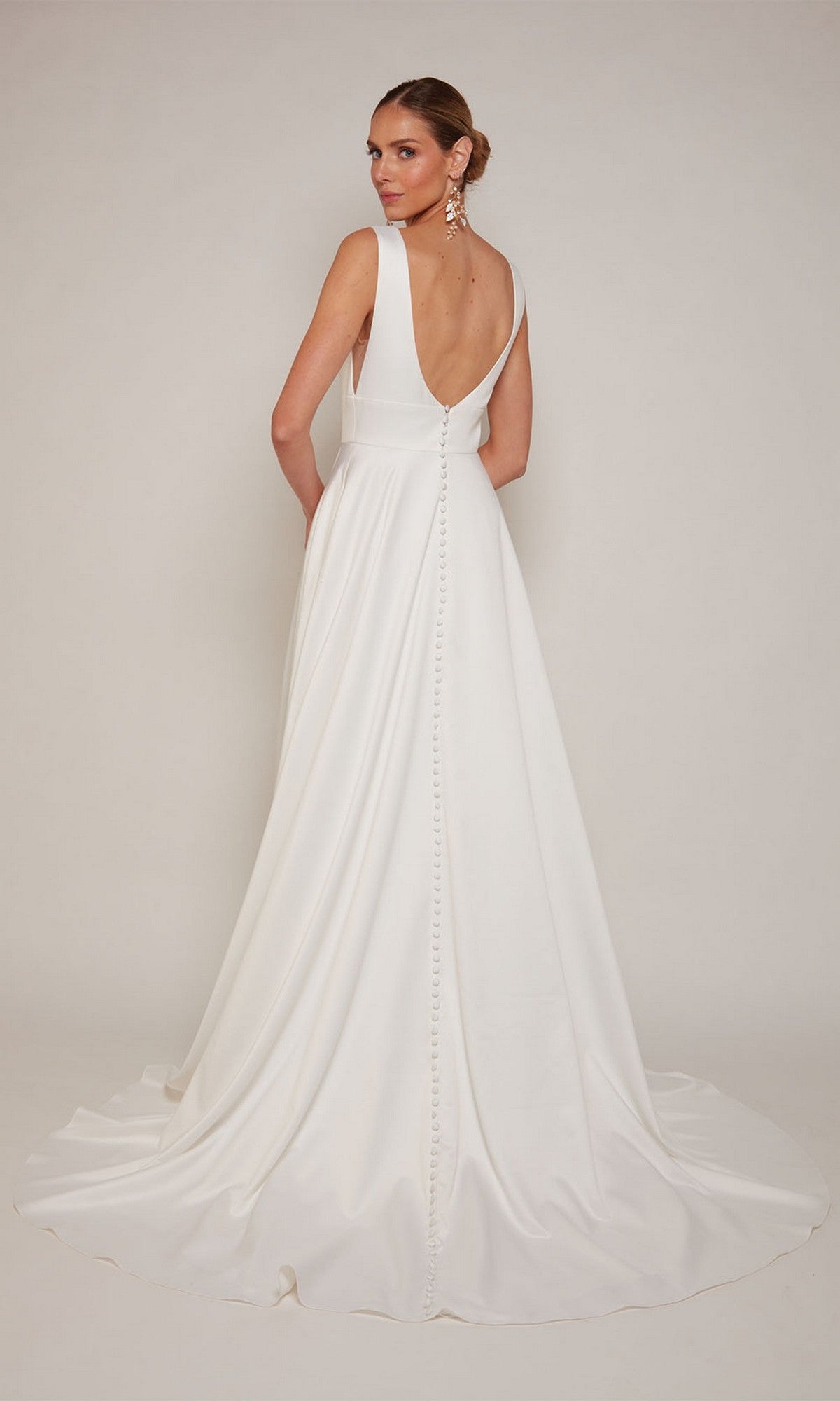 A woman models the Alyce 7096, a long formal white satin dress featuring a graceful train and button details down the back, against a plain background.